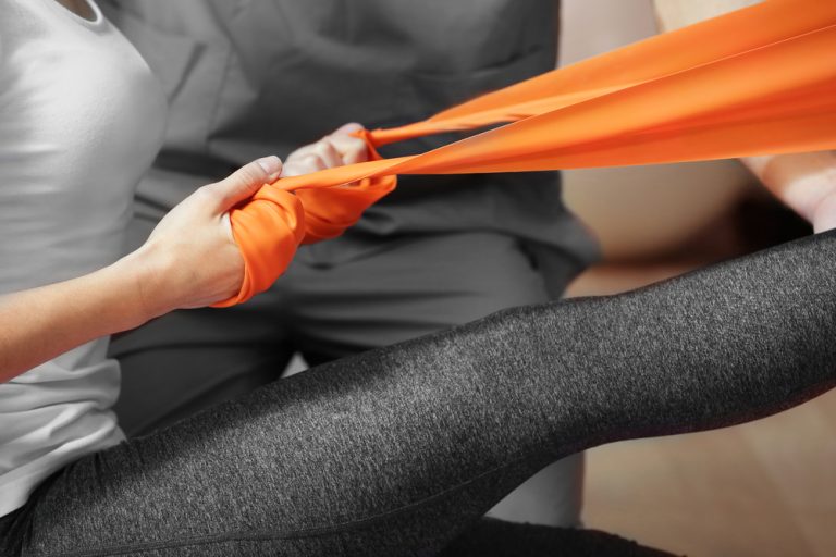 12-11-Gedragsmatige interventies verbeteren oefentrouw bij fysiotherapie_576822754 Jonge dame doet oefeningen bij de fysiotherapeut met kleurige elastische band.