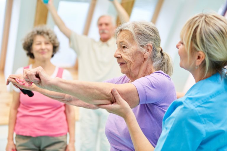 13-19-De bijwerkingen van oefentherapie_1166863978 Oudere dame die onder begeleiding van fysiotherapeute veilig oefent.