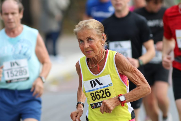 Oudere vrouw in gele hardloopoutfit rent enthousiast tijdens een marathon, omringd door andere deelnemers