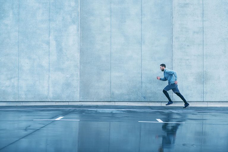 Man with sports clothes runs fast next to concrete wall Male runner sprints next to big concrete wall. He wears sports clothes. It has been raining and the street is wet.