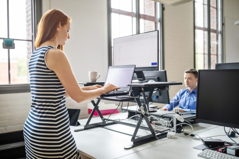 Businesswoman working at ergonomic standing desk Business woman working on laptop computer at ergonomic standing desk. Female professional working at her desk with male colleague working at the back.