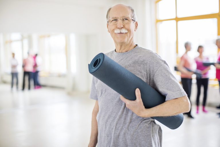 Senior man holding exercise mat in yoga class Confident senior man standing in yoga class. Portrait of smiling elderly male holding rolled up exercise mat. He is wearing sports clothing.