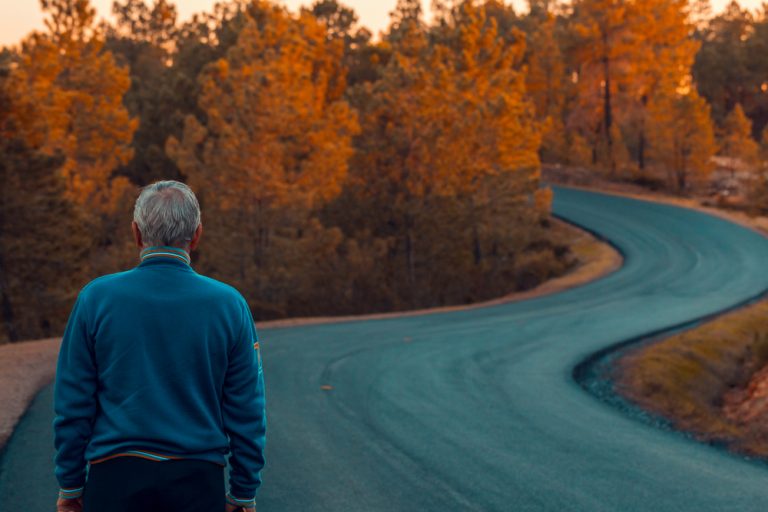 Active Active senior man stands alone on lonely road between mountains. Older man of back walking on lonely highway