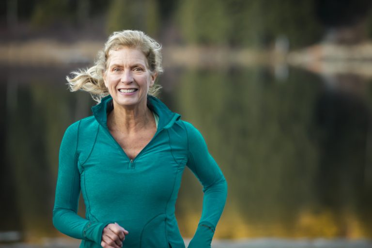 Close up of senior woman running by lake. Mature woman with long blond hair in her 60’s running around lake.
