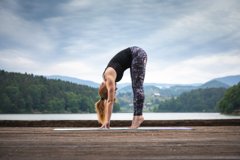 Woman Doing Yoga By The Lake Woman doing yoga on the wooden jetty. Standing Forward Bend.