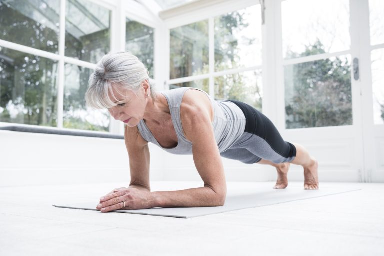 Senior woman in plank position in conservatory Woman in her 60s doing pilates at home on a yoga mat. She is balancing on her forearms and looking down towards the floor.