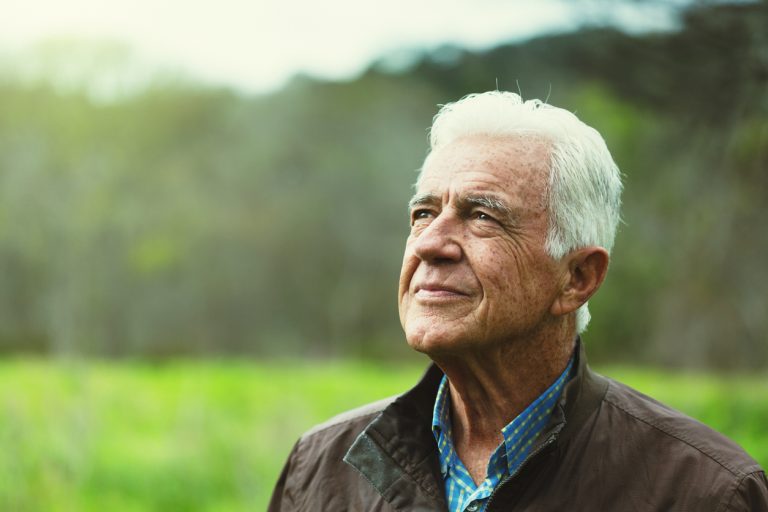 Smiling gently A white-haired senior man walking in the green countryside looks upwards smiling wistfully.