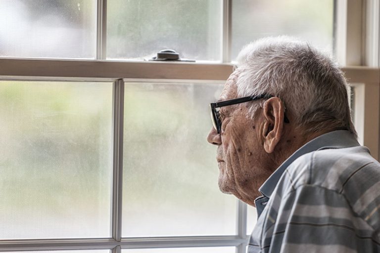 A hearing impaired elderly senior adult man wearing a hearing aid is sitting staring through the hazy, speckled, unwashed window and grungy mesh screen of a living room window at home. He has early stage dementia and will sit motionless for minutes at a time gazing engrossed at the outside world.