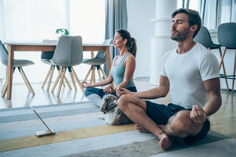 Young couple meditating in Lotus position at home. Beautiful young couple and their cute pet dog sitting on the floor and practicing yoga. Sporty couple using digital tablet for online yoga lessons.
