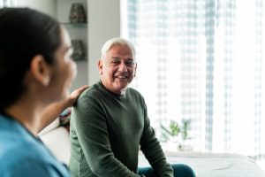 Oudere man glimlacht tijdens een gesprek met een zorgverlener die hem bemoedigend op de schouder raakt.