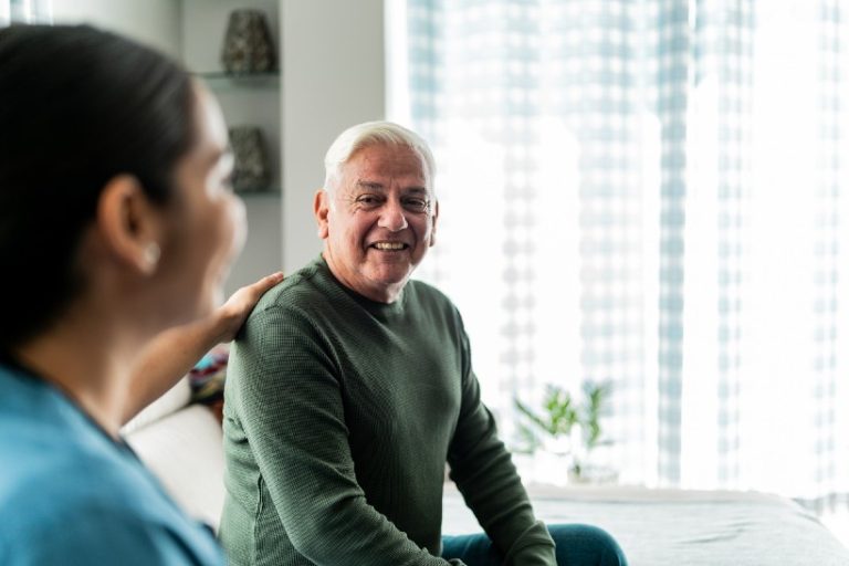 Oudere man glimlacht tijdens een gesprek met een zorgverlener die hem bemoedigend op de schouder raakt.
