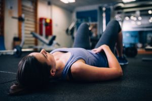 Een jonge vrouw ligt op haar rug op de vloer van een fitnessruimte, in sportkleding, met haar handen rustend op haar buik. De ontspannen houding illustreert het thema van relaxatietraining bij chronische nekpijn, zoals besproken in het artikel van Psychfysio.
