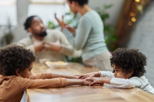 Twee kinderen zitten stil aan een tafel terwijl op de achtergrond hun ouders in een verhitte discussie verwikkeld zijn. Het beeld illustreert de emotionele impact van ouderlijke conflicten, een thema dat in het artikel over de link tussen scheiding en het ontstaan van chronische pijn wordt besproken.