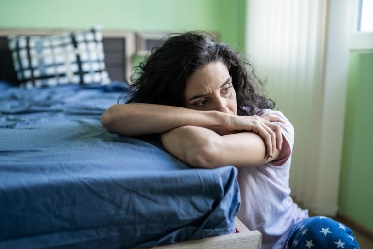 Vrouw zit op de grond naast haar bed en kijkt verdrietig voor zich uit, symbool voor het emotionele verlies en de rouw bij chronische pijn.