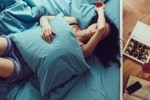 Vrouw ligt vermoeid in bed met een kussen over haar buik; naast het bed liggen chocolade en een glas drank — symbolisch voor comfort eten bij pijn en stress.