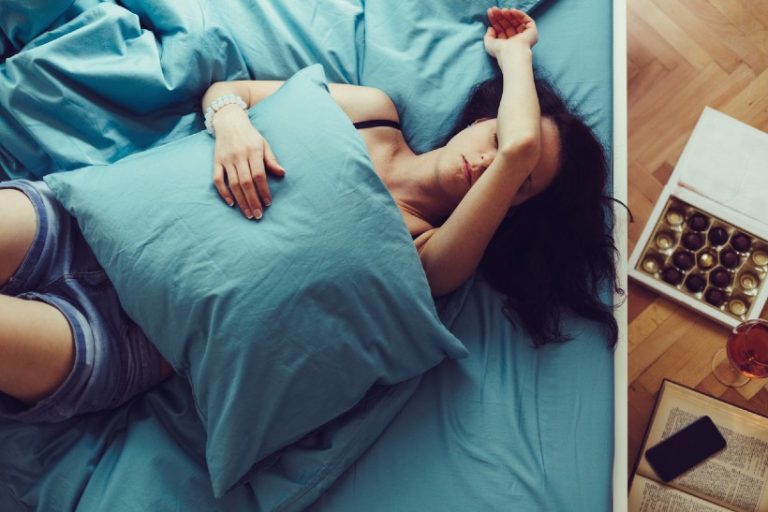 Vrouw ligt vermoeid in bed met een kussen over haar buik; naast het bed liggen chocolade en een glas drank — symbolisch voor comfort eten bij pijn en stress.