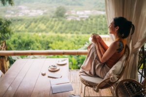 Een vrouw zit ontspannen op een veranda met uitzicht op een groen, heuvelachtig landschap. Ze houdt een kop thee vast en kijkt naar buiten, terwijl op de houten tafel voor haar een notitieboek, pen en een bordje met gebak liggen. Het beeld straalt rust en mindfulness uit, passend bij het thema van pijneducatie en mindful bewegen bij chronische lage rugpijn.