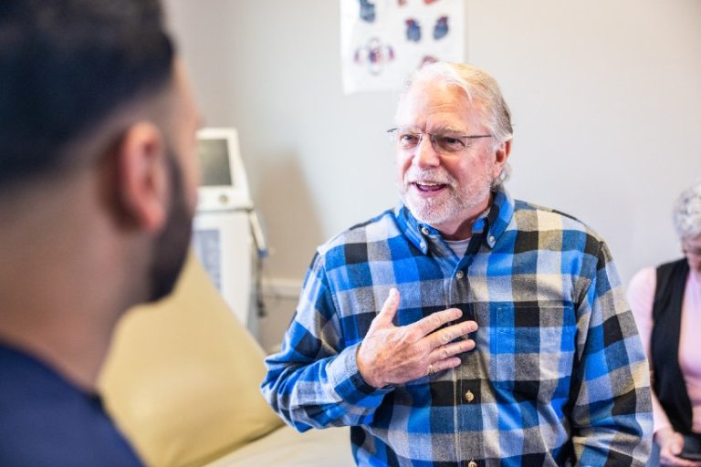 19-18-Het bevragen van de psychosomatische competentie van de patiënt-iStock-2205398583 Een oudere man met grijs haar en een bril zit in gesprek met een zorgverlener in een klinische setting. De man heeft zijn hand op zijn borst gelegd terwijl hij iets uitlegt, wat duidt op een open en persoonlijke uitwisseling.