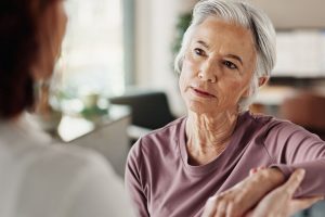 Een oudere vrouw met grijs haar luistert aandachtig tijdens een gesprek in een rustige binnenruimte. Haar gezichtsuitdrukking toont concentratie en lichte emotie, passend bij het thema van het artikel over hoe mensen met chronische pijn minder goed onderscheid maken tussen pijn en emoties.