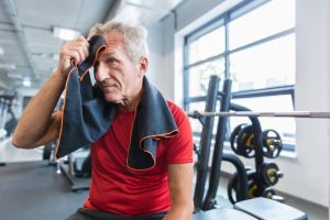 Oudere man rust uit na een training in de fitnessruimte; hij veegt zijn gezicht af met een handdoek. De foto illustreert hoe kracht- en duurtraining op latere leeftijd elkaar kunnen versterken.