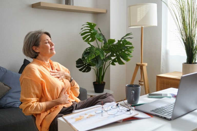 Vrouw van middelbare leeftijd zit ontspannen thuis met handen op borst en buik en oefent rustige ademhaling achter haar laptop, passend bij behandeling van fibromyalgie en milde chronische hyperventilatie.