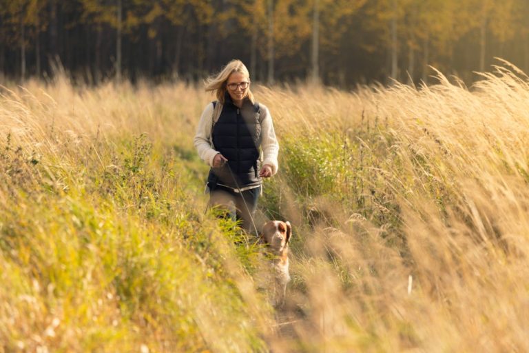 Vrouw wandelt met hond door hoog gras in herfstlicht, rustige buitenomgeving.