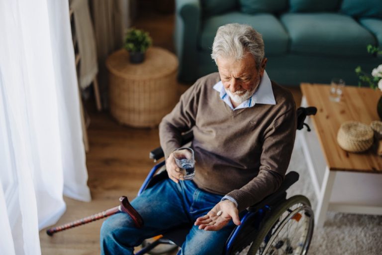 Oudere man in een rolstoel houdt een glas water en medicijnen in zijn hand in de woonkamer.