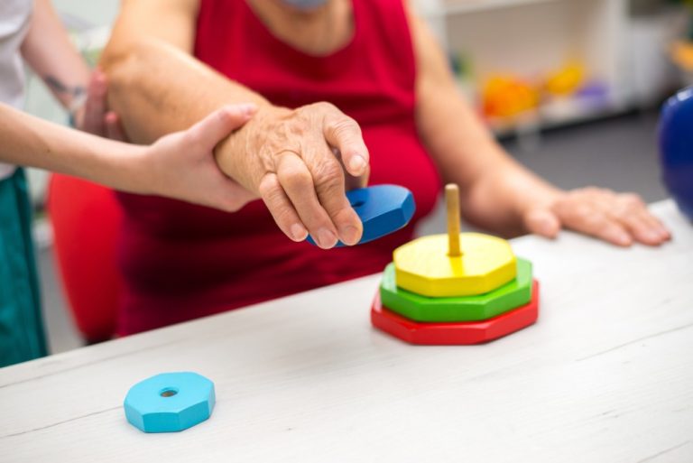 Revalidatie na CVA: begeleider ondersteunt hand-arm oefening met stapelringen op tafel.