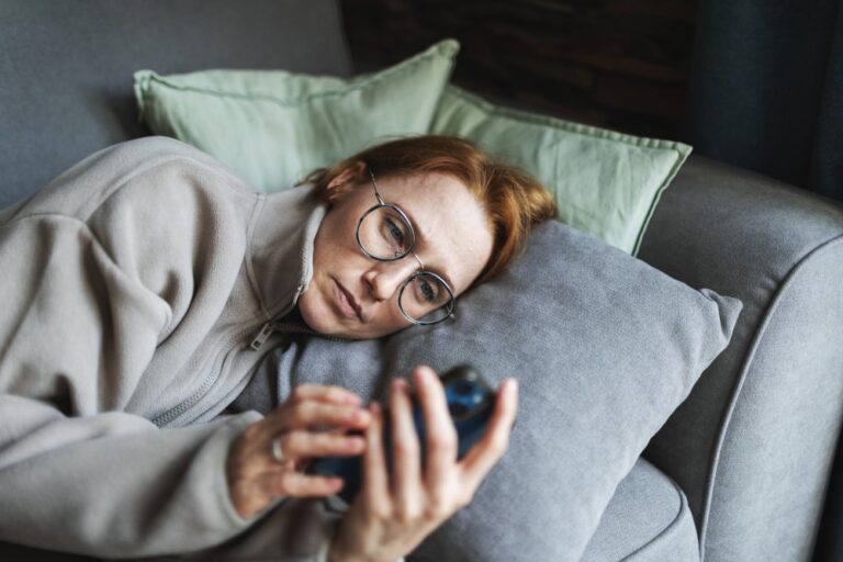 Vrouw ligt op de bank en kijkt somber naar haar smartphone, passend bij thema chronische pijn en psychosociale factoren.