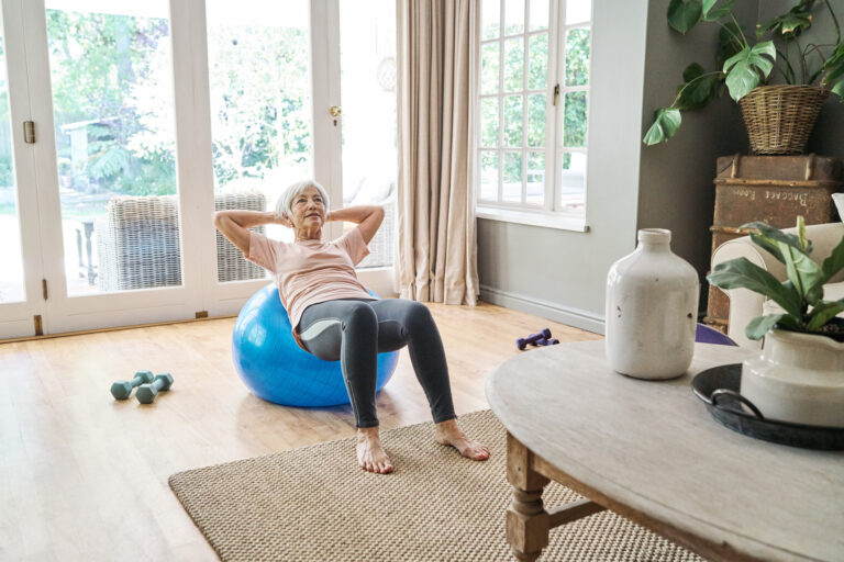 Oudere vrouw doet buikspieroefeningen op een grote oefenbal in de woonkamer, met halters op de vloer.