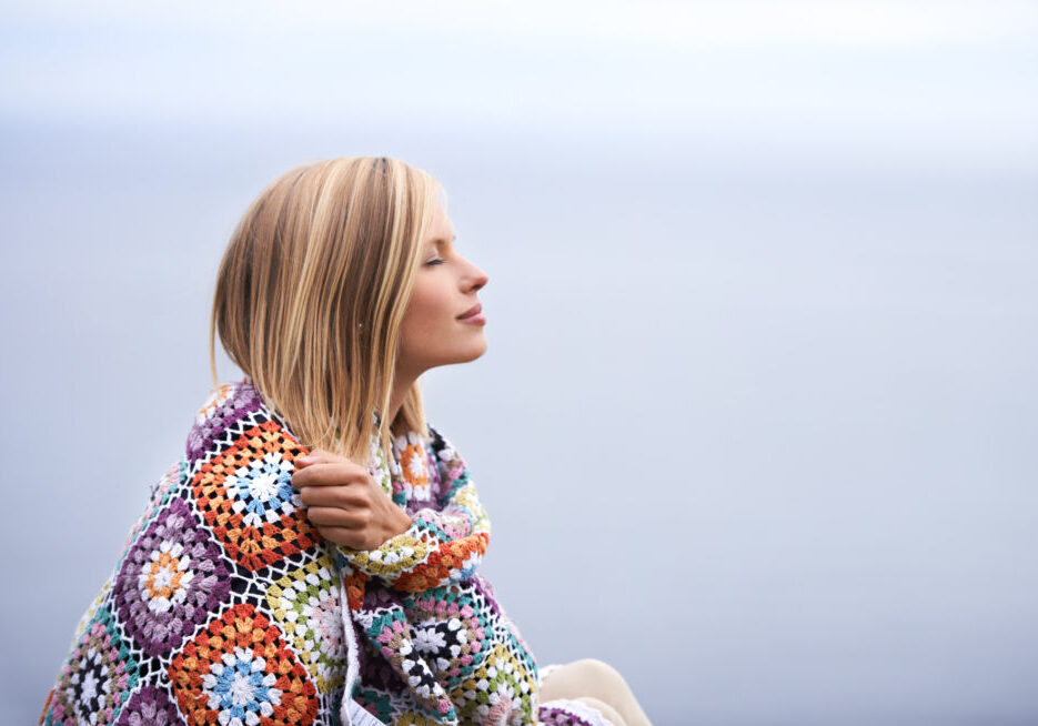Taking in the morning Shot of a beautiful young woman wrapped in a blanket sitting at the ocean