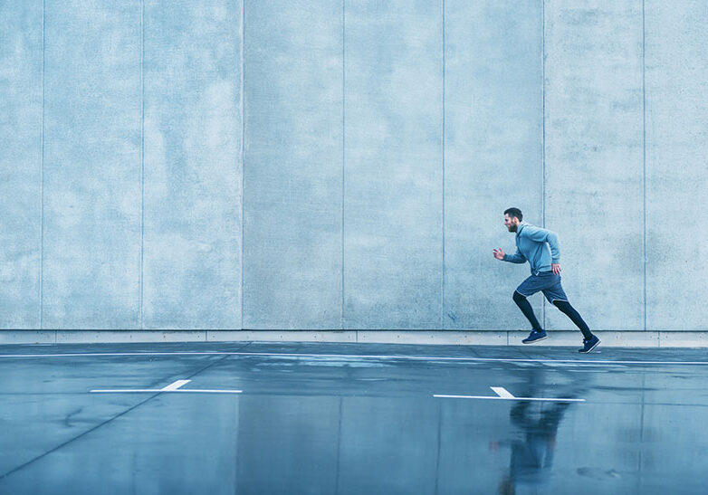 Male runner sprints next to big concrete wall. He wears sports clothes. It has been raining and the street is wet.