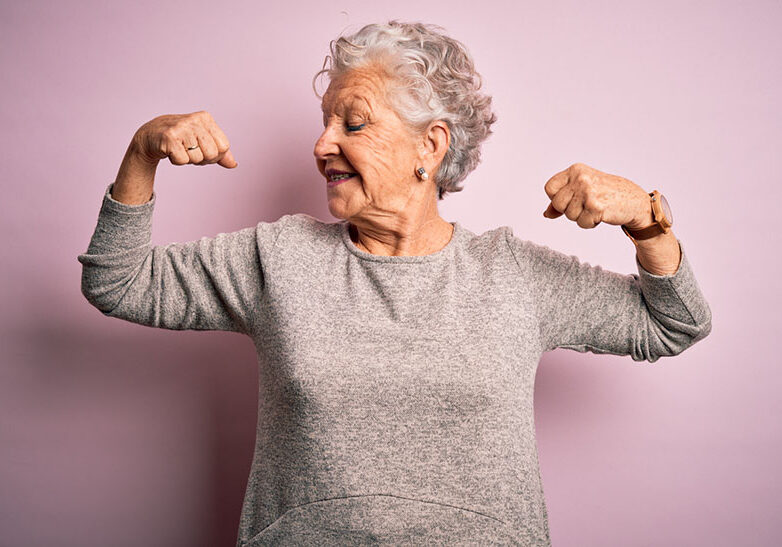 Senior beautiful woman wearing casual t-shirt standing over isolated pink background showing arms muscles smiling proud. Fitness concept.