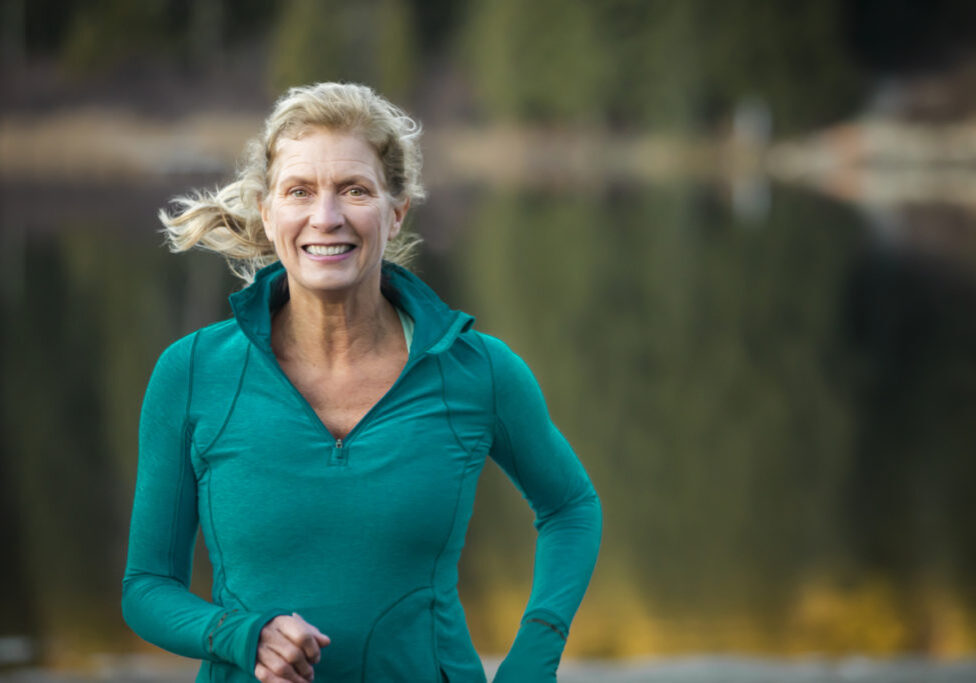 Mature woman with long blond hair in her 60’s running around lake.