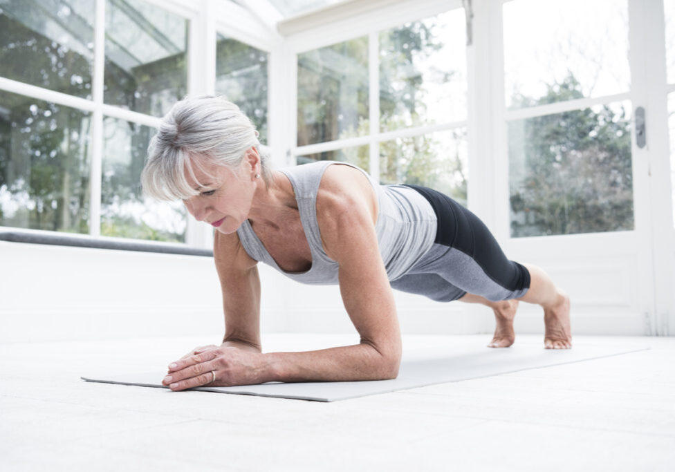 Woman in her 60s doing pilates at home on a yoga mat. She is balancing on her forearms and looking down towards the floor.