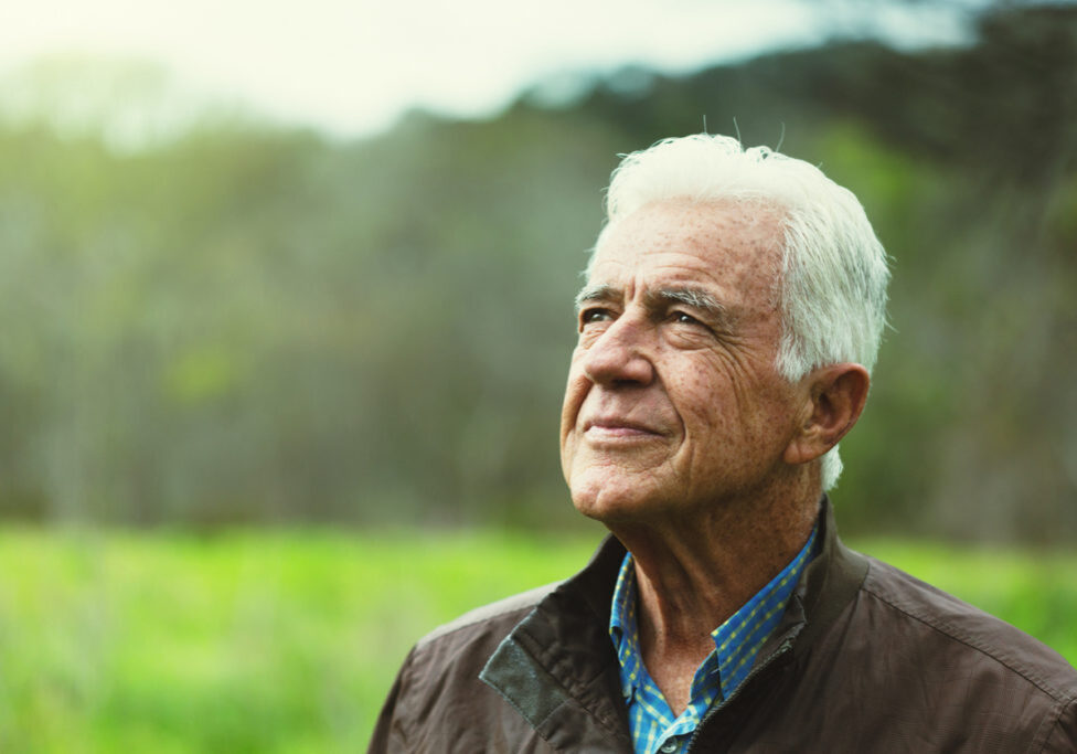 A white-haired senior man walking in the green countryside looks upwards smiling wistfully.