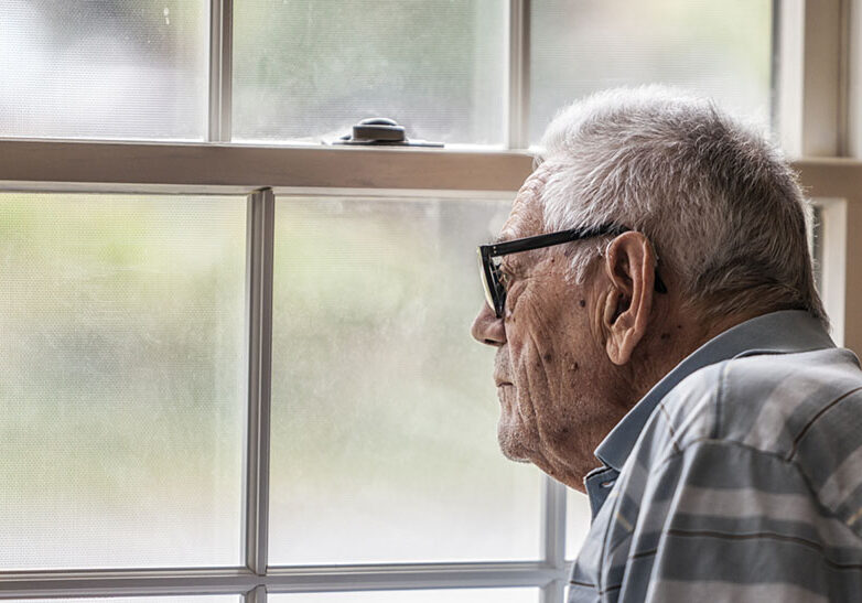 A hearing impaired elderly senior adult man wearing a hearing aid is sitting staring through the hazy, speckled, unwashed window and grungy mesh screen of a living room window at home. He has early stage dementia and will sit motionless for minutes at a time gazing engrossed at the outside world.