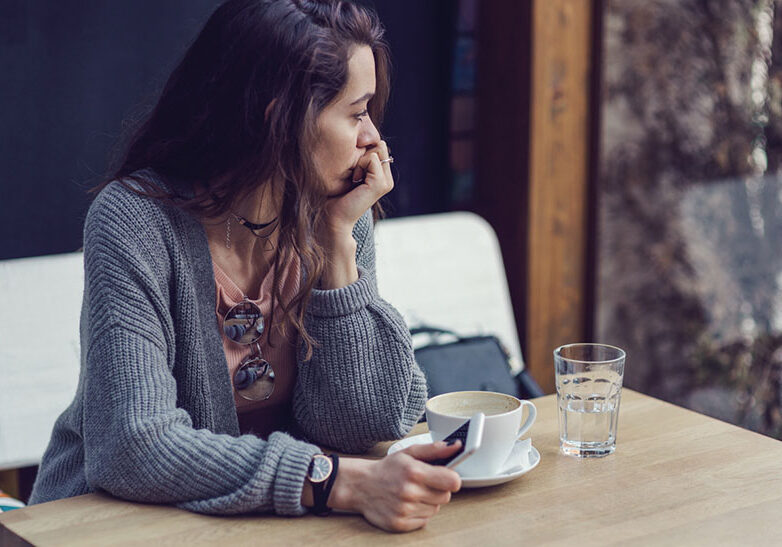Brunette female holding her mobile phone, looking through the window and drinking coffee