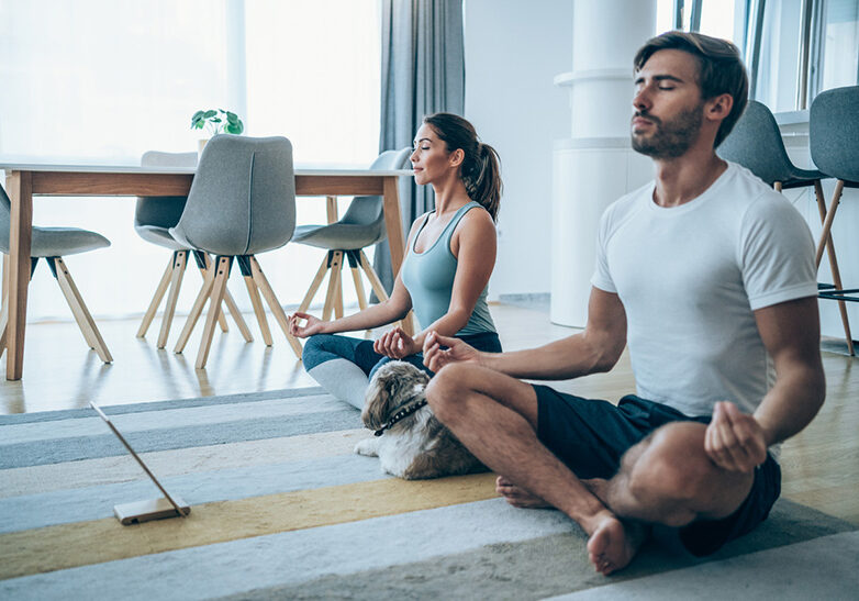 Beautiful young couple and their cute pet dog sitting on the floor and practicing yoga. Sporty couple using digital tablet for online yoga lessons.